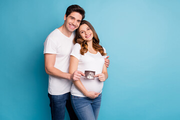 Poster - Photo of young cheerful positive smiling happy family hold ultrasound photo of baby isolated on blue color background