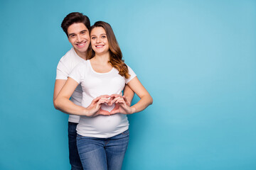 Poster - Photo of young cheerful positive smiling couple pregnant wife and husband showing heart on tummy isolated on blue color background