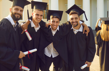 Wall Mural - Group of smiling men students university graduates standing, hugging and celebrating getting diplomas with classmates and university building at background. Successful univesity graduation concept