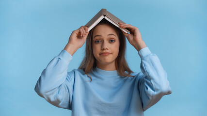 Wall Mural - teenage girl in soft sweatshirt holding book above head isolated on blue