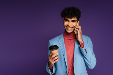 Wall Mural - cheerful african american man in blue blazer talking on smartphone while holding paper cup on purple