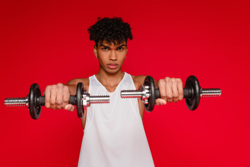 Wall Mural - sportive african american man in tank top exercising with dumbbells on blurred foreground isolated on red