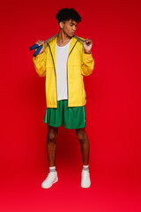 Wall Mural - full length of sportive african american man in yellow rain jacket standing with skipping rope on red