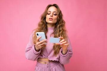 Beautiful young blonde curly woman wearing pink clothes isolated over pink background using mobile phone paying online shopping through credit card looking at smartphone display