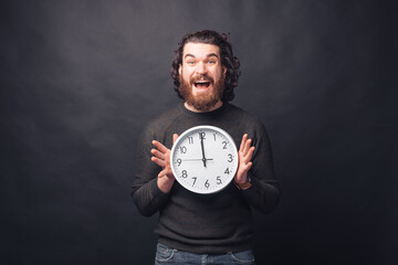 Wall Mural - Portrait of amazed bearded man holding clock at twelve a clock over black background.