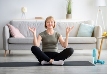 Wall Mural - Balanced senior woman sitting in lotus pose and making gyan mudra with her hands, meditating on sports mat at home