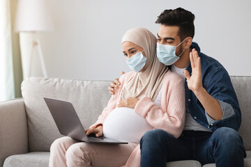 Wall Mural - Pregnant muslim couple wearing medical masks making video call with laptop at home