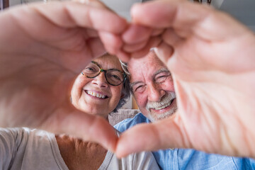 Couple of two old and happy seniors having fun at home on the sofa doing a heart shape with their hands and fingers looking at the camera. 