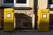 Zwei gelbe Briefkästen zwischen Sonne und Schatten stehen dicht vor einer Hausfassade. Ein Teil Fenster und Abflussrohr ist zu sehen.