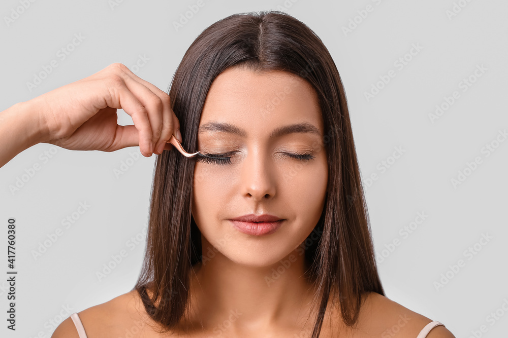 Beautiful young woman applying fake eyelashes against light background