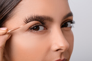 Poster - Beautiful young woman applying fake eyelashes against light background, closeup