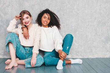 Two young beautiful smiling international hipster female in trendy summer jeans clothes. Sexy carefree women posing near gray wall in studio. Positive models having fun. Concept of friendship