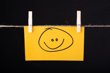 A smile Emoji Drawn on a Yellow color sticky note hanging with a wire on black background