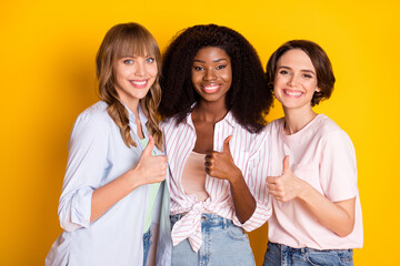 Wall Mural - Portrait of three attractive cheerful girls embracing showing thumbup advert isolated over bright yellow color background