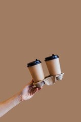 A hand holds two cups of coffee on a stand on a brown background. Cappuccino with almond milk to take away. Hot coffee in a craft cup. Space for text and logo. Mockup cup of coffee