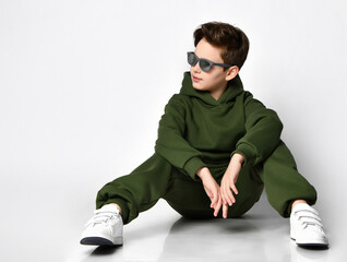 Cool schoolboy in a green warm fleece tracksuit and sunglasses sitting on a gray background. Child looks away at a free space for text. Concept of children's sportswear and children's style.