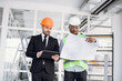 © Sofiia Shunkina - International business partners in formal suit and helmets doing drawings inspection. Two multiracial men standing indoors and holding blueprint with digital table.
