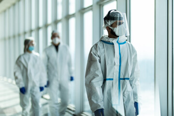 Wall Mural - Three disinfectors or doctors walk in protective uniform walking in hospital corridor. Scientists in biohazmat suits walking along chemical laboratory hallway