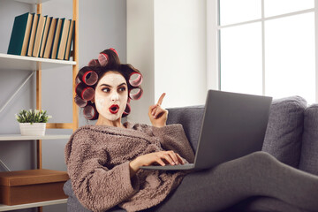 Surprised woman points finger up as she finds amazing idea online. Happy lady in curlers and face mask sitting on sofa, browsing Internet blog on laptop and learning good life hacks and practical tips