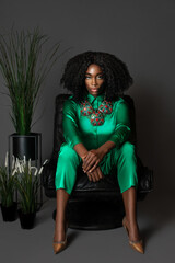 Portrait of a sensual black woman with curly long black hair and beautiful makeup sitting in a leather seat by herself in a studio with a grey background wearing a green jumpsuit and colorful jewelry.