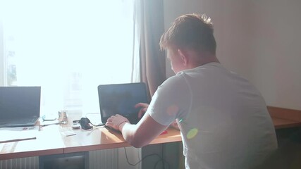 Fototapete - The guy uses the laptop keyboard at the table. He does study assignments. Sunlight outside the window. Camera movement.