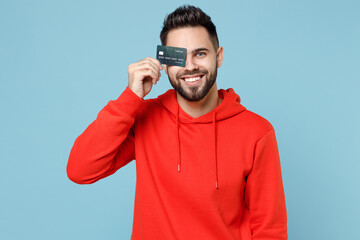 Canvas Print - Young caucasian smiling happy attractive satisfied rich bearded student man 20s wearing casual red orange hoodie covering eye with credit bank card isolated on blue color background studio portrait.