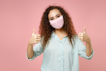 Wall Mural - Young black african curly woman in blue shirt reusable face mask to safe from coronavirus virus covid-19, pandemic quarantine show thumb up gesture isolated on pastel pink background studio portrait