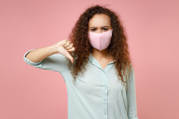 Wall Mural - Young black african curly woman in blue shirt reusable face mask safe from coronavirus virus covid-19, pandemic quarantine show thumb down gesture isolated on pastel pink background studio portrait