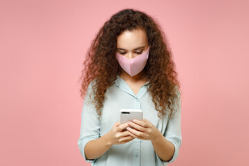 Wall Mural - Young black african woman in blue shirt face mask from coronavirus covid-19 pandemic quarantine using mobile cell phone browsing symptoms read news isolated on pastel pink background studio portrait.