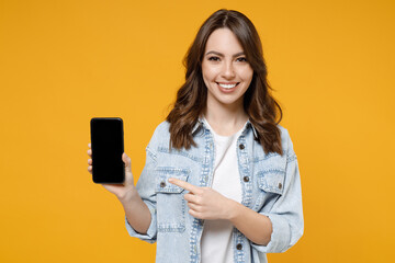 Wall Mural - Young smiling happy fun woman 20s wear stylish casual denim shirt white t-shirt point index finger on mobile cell phone with blank screen workspace area isolated on yellow background studio portrait