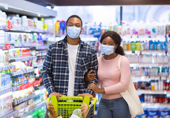 Wall Mural - Black couple in masks doing grocery shopping together during covid lockdown, walking with full trolley at supermarket