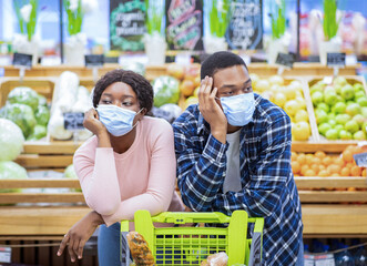 Wall Mural - Bored millennial couple in face masks leaning on shopping cart, sick and tired of making purchases at mall