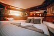 A wide-angle fragment of an interior of a luxurious boat cabin with a big neatly made bed, lacquered wood paneling decoration, stripe of mirrors, and an area with portholes with a sunny day outside
