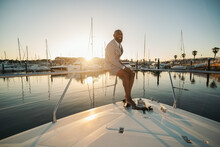 A Cheerful Elegant Mature Bald Bearded Black Man In A Summer Suit Consisting Of Striped Shorts And Blazer Is Sitting On The Railings Of His Luxurious Yacht, Laughing And Enjoying The Dramatic Sunset