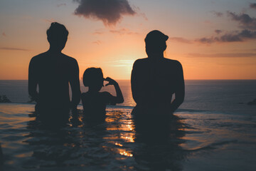 Silhouette family, including father, mother and daughter stands by the swimming pool and enjoying a beautiful sunset view over the sea.