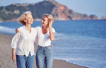 Outdoor portrait of smiling happy caucasian senior mother with her adult daughter hugging and walking on sea beach.