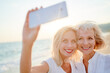 © luengo_ua - Outdoor portrait of smiling happy caucasian senior mother with her adult daughter taking selfie on smartphone on sea beach.