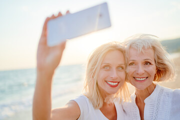 Outdoor portrait of smiling happy caucasian senior mother with her adult daughter taking selfie on smartphone on sea beach.