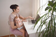 © Алина Троева - Young mother work studying from home with laptop computer during quarantine, little cute toddler baby on lap. Home office, parenthood. Remote work, distance education. Video conference virtual event.