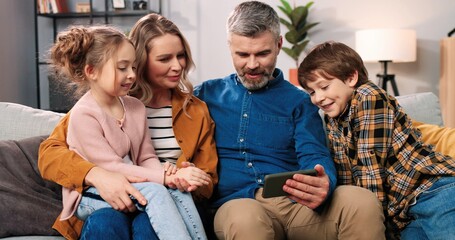 Wall Mural - Portrait of cheerful happy Caucasian family small cute kids with caring parents sitting on sofa in room in apartment talking watching cartoon at cellphone screen, video chatting, using digital gadget