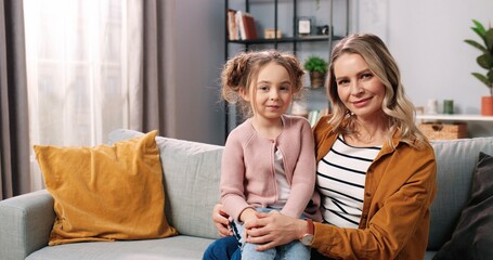 Wall Mural - Portrait of joyful lovely Caucasian little adorable girl sitting on couch in living room with pretty mother looking at camera and smiling. Caring mom with kid spending time together, family concept