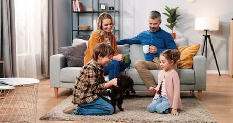 Wall Mural - Caucasian lovely happy family gathered at home together. Mother and father sitting on sofa and drinking coffee watching their little kids girl and boy playing with cute animal puppy, home pet concept