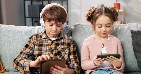 Wall Mural - Close up of Caucasian little children brother and sister sitting on sofa in living room using gadgets. Small girl watching cartoon on smartphone while kid boy playing game on tablet, leisure concept