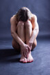 A naked woman sits on the floor with her head down. Dark background