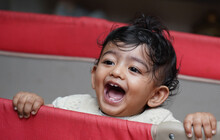 A Closeup Phot Of An Adorable Indian Toddler Baby Boy Smiling With Dimple In Cheeks And Standing Inside A Playpen.