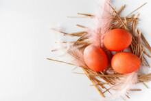 Orange Easter Eggs And Feathers In A Nest On A White Background.