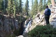 Female hiker with braids, backpack and hiking poles stands near Kings Creek Falls in Lassen Volcanic National Park. Hiker in silhouette and shade with water fall cascade in full light. 