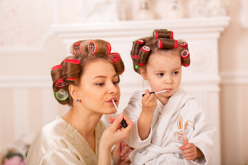 Adorable little girl with her mother in hair curlers apply makeup. Mom teaches daughter to use cosmetics. Beauty day. Girls are such girls