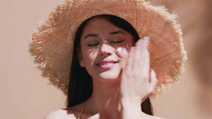 Wall Mural - Young attractive asian woman in straw hat shows a drop of spf cream on her finger and applies it to her face, looks at the sun then at camera and smiles on beige background with palm tree leaf shadows