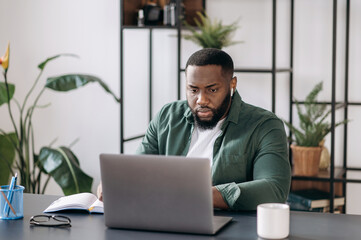 Concentrated serious young successful multiracial black ceo, programmer or freelancer working using laptop while sitting at his work desk